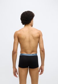 Cuecas boxer pretas com uma cintura azul-claro marcada com "HUGO", apresentando um ajuste justo e tecido suave. Modelo visto de costas.