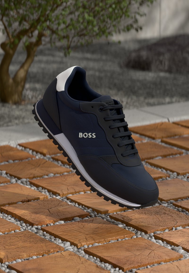 Pantofii sport navy cu accente negre și albe, material texturat, design cu șireturi și talpă robustă, expuși deasupra unei suprafețe de plăci cu model.