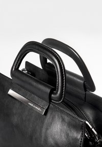 Musta nahkainen käsilaukku, jossa on kaksi pyöreää kahvaa, vetoketjukiinnitys ja metallinen korostelappu, jossa on brändin logo. Tasainen pinta, jossa on hillitty tikkaus.