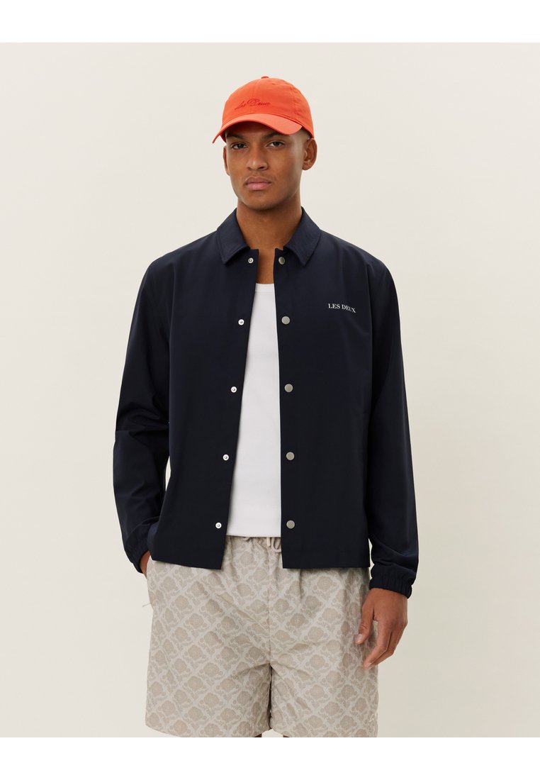 Navy button-up jacket with a collar and silver snap buttons, worn over a white tank top and patterned light shorts, paired with an orange cap.