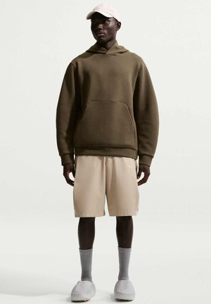 Man staat, draagt een olijfgroene hoodie, beige korte broek, grijze sokken, witte sneakers en een lichtbeige baseballpet op een effen achtergrond.