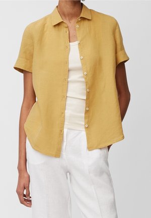 Person wearing a short-sleeve mustard yellow button-up shirt over a white tank top and white pants, standing with hands relaxed.