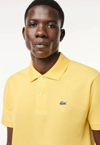 Polo amarillo hecho de tejido texturizado con tres botones y un pequeño logo bordado de cocodrilo verde en el lado izquierdo del pecho.