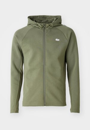 Groene ritshoodie met een capuchon, gemaakt van gladde stof. Heeft raglanmouwen en een klein logo op de borst. Textuur panelen aan de zijkanten.