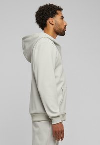Lys grå zip-up hoodie, blødt stof, kängurulomme, elastiske manchetter og kant, med en snoretrækshætte, sideview af mandlig model.