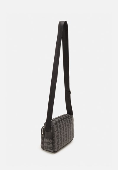 Sac à bandoulière noir, rectangulaire, avec un motif géométrique, fermeture à zip et une longue sangle ajustable en tissu robuste. Détail de surface texturée.