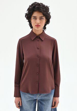 Blusa marrone a maniche lunghe con scollo con collo, chiusura con bottoni e tessuto morbido e liscio; abbinata a jeans di denim chiaro.