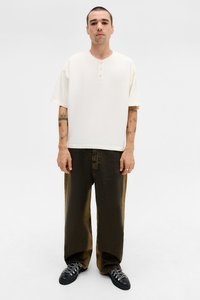 Off-white short-sleeve henley shirt with three buttons, paired with loose olive-green denim pants and black lace-up shoes.