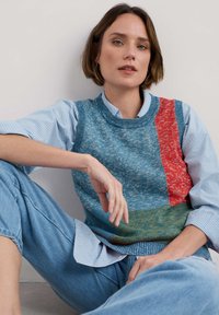 Gestrickter ärmelloser Pullover mit blauen, roten und grünen Abschnitten sowie einem strukturierten Muster. Das Model trägt ein gestreiftes Hemd mit Kragen darunter.
