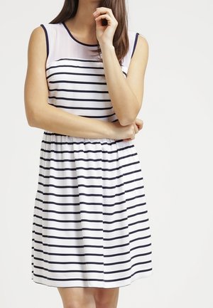 Woman wearing a sleeveless white dress with horizontal black stripes and sheer upper chest panel, standing with arms crossed against a white background.