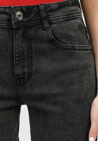 Gros plan sur la ceinture en denim noir avec un bouton métallique et une poche avant, portée par une personne dont une partie de la main est visible.