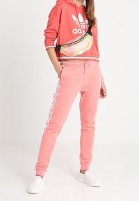 Korallfärgad hoodie med vit Adidas-logotyp och randiga ärmar, kombinerad med matchande korallfärgade joggers med vita sidostripes. Vita slip-on skor och en genomskinlig midjeväska.