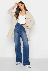 Beige übergroßes Hemd mit Knöpfen, weißes Tanktop, hoch taillierte blaue Schlagjeans und weiße Sneaker, mit glattem Material und lässigem Design.