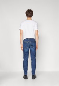 Jeans de mezclilla azul con corte slim, que presentan bolsillos traseros y costuras amarillas, combinados con una camiseta blanca sencilla y zapatos negros.