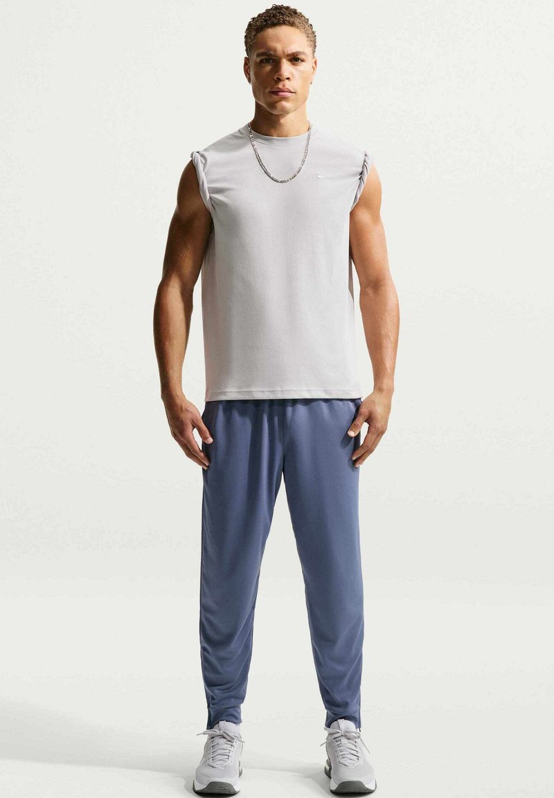 Chemise grise sans manches avec un col rond, associée à un pantalon de sport bleu. Le mannequin porte une chaîne en argent et des baskets grises.