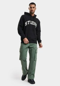 Svart hettegenser med teksten "STUD", kombinert med grønne cargo-bukser med sidelommer og sølvfester, brukt med svarte sneakers.