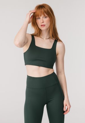 Žena s červenými vlasmi nosiaca tmavozelenú športovú podprsenku s hranatým výstrihom a legíny s vysokým pásom, pózujúca na svetlošedom pozadí s rukou pri vlasoch.
