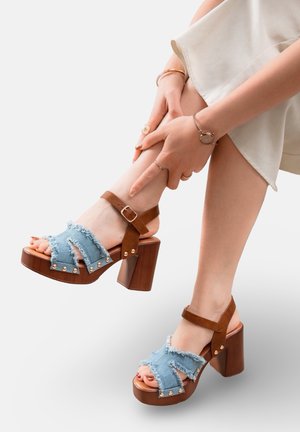 Femme portant des sandales à talons hauts marron avec des brides en jean délavé, touchant sa cheville, vêtue d'une jupe de couleur claire.