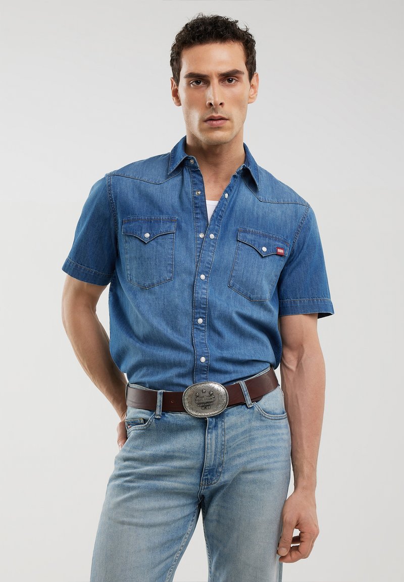 Camicia in denim a maniche corte di colore blu chiaro, con due tasche frontali, bottoni a pressione e colletto a punta, abbinata a jeans blu sbiaditi e una cintura marrone.
