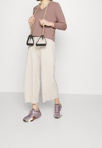 Maglietta beige a scollo a V, pantaloni larghi color crema con texture e scarpe da ginnastica viola; tiene in mano i manici di una banda di resistenza nera.