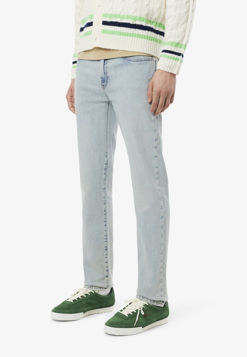Jeans in denim azzurro chiaro con una vestibilità slim, abbinati a un cardigan in maglia cable-knit color crema con strisce verdi e blu navy e scarpe da ginnastica verdi.