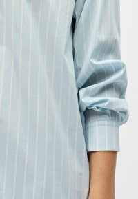 Chemise à rayures bleu clair à manches longues ; tissu lisse avec un léger éclat ; présente des poignets retroussés et des rayures verticales pour la texture.