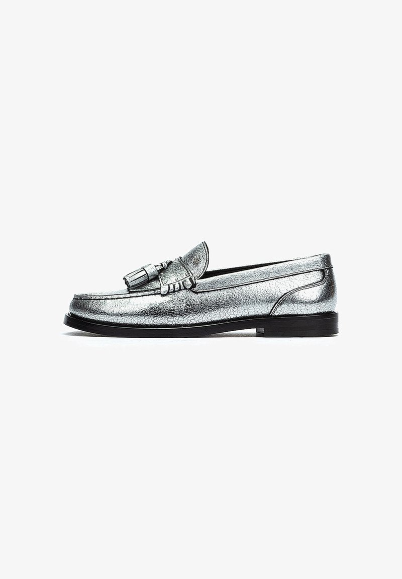 Mocasines plateados metálicos con un acabado texturizado, que presentan un detalle de borlas en el frente, puntera redonda y una suela negra baja.