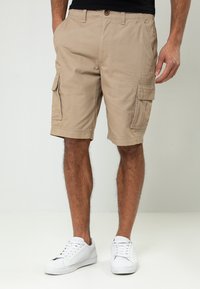 Homme portant un short cargo beige avec des poches latérales et des baskets blanches, debout devant un fond clair uni.