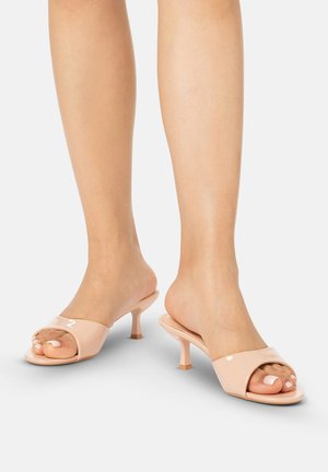 Voeten met lichtroze open-teen kittenhielmuiltjes met een gladde afwerking op een effen lichte achtergrond.