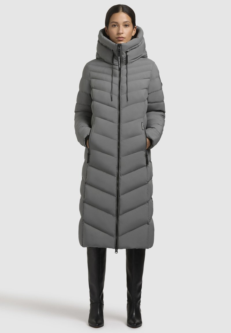 Grijze lange pufferjas met een hoge kraag, voorzien van een rits aan de voorkant, chevron-quilting en zijzakken. Draagt met zwarte kniehoge laarzen.