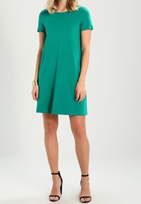 Vrouw in een groen jurk tot op de knie met korte mouwen, zwarte enkelsandalen met hoge hakken en een gouden armband om haar linkerpols.