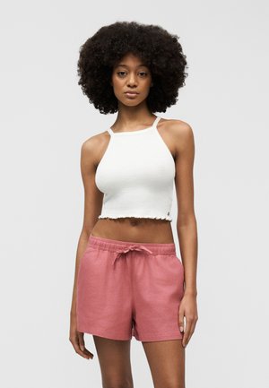 Fiatal nő göndör hajjal, fehér ujjatlan haspólót és rózsaszín zsinóros rövidnadrágot viselve, egyszínű háttér előtt áll.