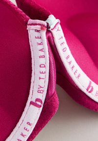 Sutien roz căptușit, cu material neted, accente delicate de dantelă și o bandă elastică cu marcă. Designul include o strălucire subtilă și textură.