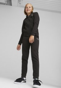 Felpa nera con zip e pantaloni jogger coordinati realizzati in tessuto morbido, con un logo bianco sul petto e sull'anca. Sneakers nere con dettagli bianchi.