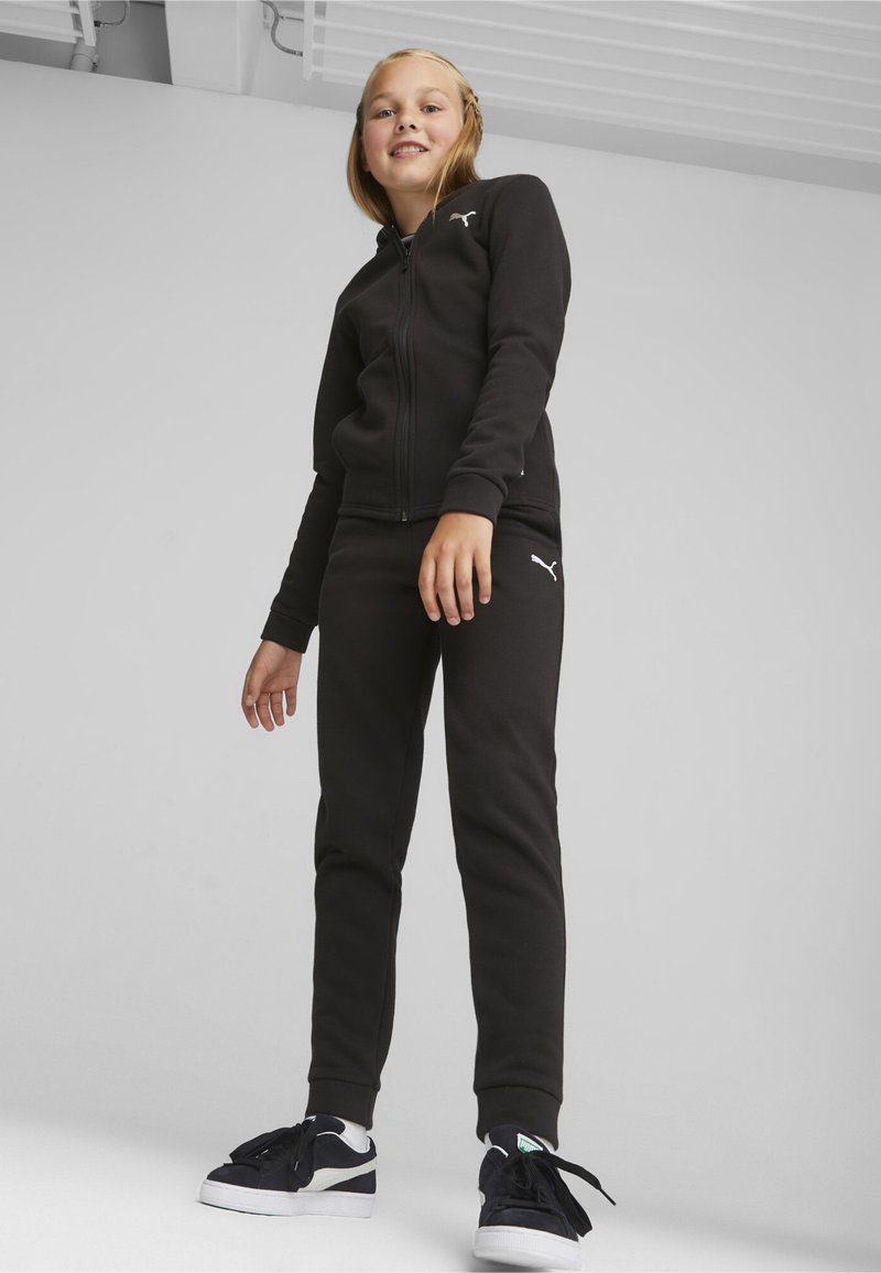 Felpa nera con zip e pantaloni jogger coordinati realizzati in tessuto morbido, con un logo bianco sul petto e sull'anca. Sneakers nere con dettagli bianchi.