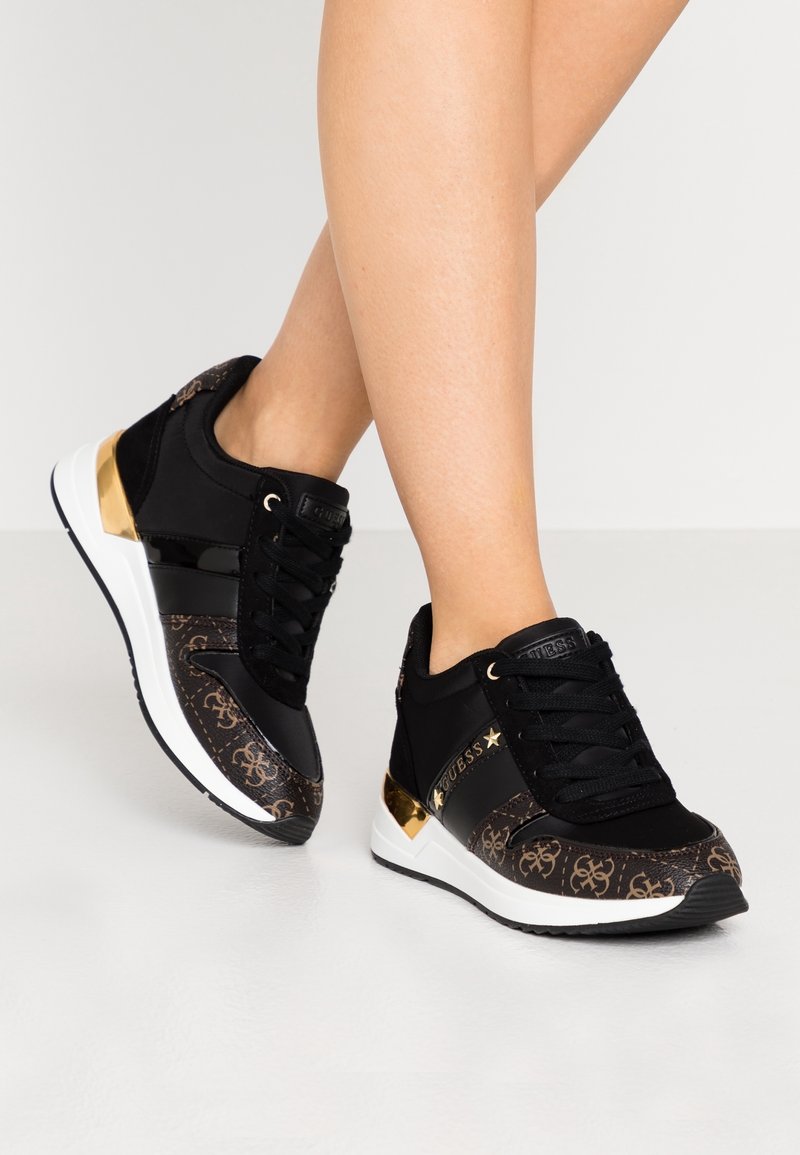 Jambes portant des baskets noires et marron avec des motifs de marque, des accents dorés, des lacets noirs et des semelles blanches sur un fond gris clair.