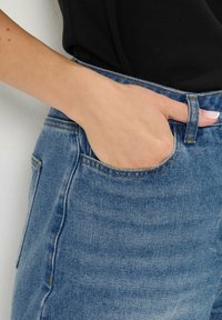 Un jean en denim bleu clair présente un design classique à cinq poches. Le tissu a une légère texture et est rehaussé de coutures orange contrastantes.