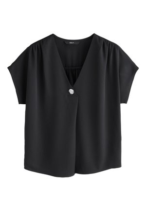 Frau geht und trägt eine schwarze V-Ausschnitt-Bluse mit einem Knopf, dunkelblaue Jeans, schwarze Slipper und minimalen Schmuck.
