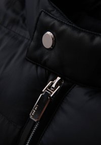 Gros plan d'une veste matelassée noire avec un bouton pression argenté et une fermeture éclair métallique. Tissu lisse et texturé avec des détails de design matelassé.