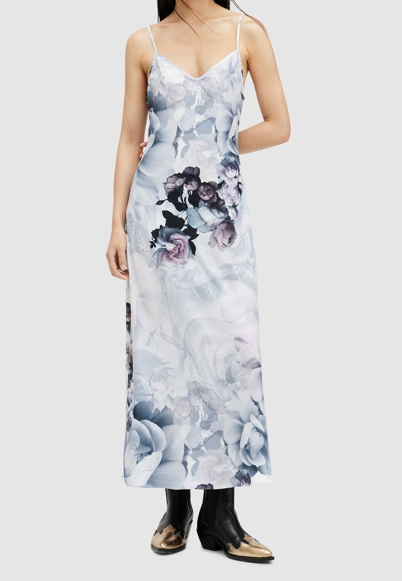 Kvinna klädd i en lång klänning med blommönster och tunna axelband, kombinerad med svarta och gyllene cowboyboots framför en enfärgad bakgrund.