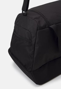 Sac de sport noir en nylon avec une texture lisse. Il dispose d'un compartiment supérieur zippé, d'une poche en filet sur le côté et d'une sangle de transport robuste.
