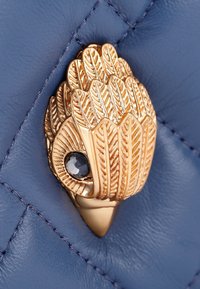Cierre decorativo de oro con patrones de hojas y una piedra preciosa oscura, sobre un fondo de cuero azul texturizado con costuras en forma de diamante.