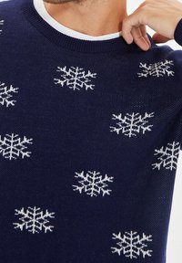 Pull marine or bleu nuit avec des motifs de flocons de neige blancs partout. Texture tricotée avec un col rond et des poignets côtelés.