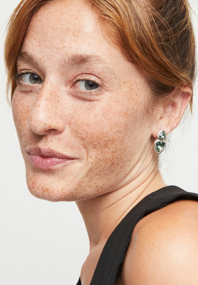 Pendientes de plata con un diseño de gema verde triangular, sobre un fondo de piel con pecas y ropa negra.