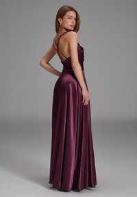 Robe de soirée longue sans manches en satin bordeaux profond. Présente un corsage ajusté avec plusieurs bretelles au dos et une jupe fluide drapée élégamment.