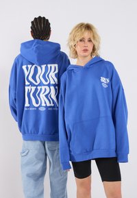 Felpa blu con il testo grafico bianco "IL TUO TURNO" sul retro e un piccolo design sul davanti. Realizzata in materiale morbido con una vestibilità ampia.