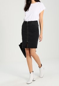 Falda lápiz de mezclilla negra con cremallera frontal, combinada con una camiseta de manga corta blanca y botines blancos, presentando un diseño minimalista.
