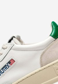 Sneaker in pelle bianca con dettaglio in camoscio beige, linguetta del tallone verde e piccolo logo della bandiera americana vicino ai lacci su sfondo semplice.