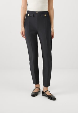 Femme portant un pantalon noir slim-tailleur avec des boutons dorés et des ballerines Mary Jane vernies noires, debout sur un fond blanc.