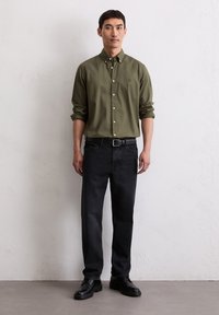 Olijfgroene button-up shirt met opgerolde mouwen, gecombineerd met zwarte denim jeans en zwarte veterschoenen, staand tegen een neutrale muur.